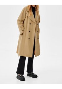Trench-coat beige à double boutonnage avec col cranté, grands boutons et poches latérales, associé à un pantalon noir évasé et des chaussures noires imposantes.