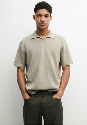 Jeune homme aux cheveux courts et bouclés portant un polo en maille beige à manches courtes et un pantalon foncé, mains dans les poches, expression neutre.