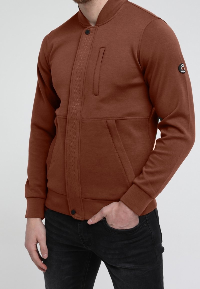 Homme portant une veste marron zippée avec une poche poitrine verticale et des poches latérales, main dans la poche d'un jean noir, fond gris.