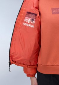 Oranžová prošívaná bunda s hladkým, lesklým povrchem. Interiér zahrnuje kapsu s grafickým prvkem hory a logem. Náplety na rukávech a lemu.