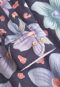 Blumenmuster im Ärmelsaum eines dunkelblauen Shirts, das große helllila und türkisfarbene Blumen zeigt, akzentuiert mit hellrosa Formen und beigen Knöpfen.