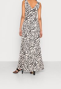 Robe maxi de couleur crème avec un motif abstrait noir, dotée d'un dos en V et d'une taille cintrée, associée à des sandales à talons hauts noires.