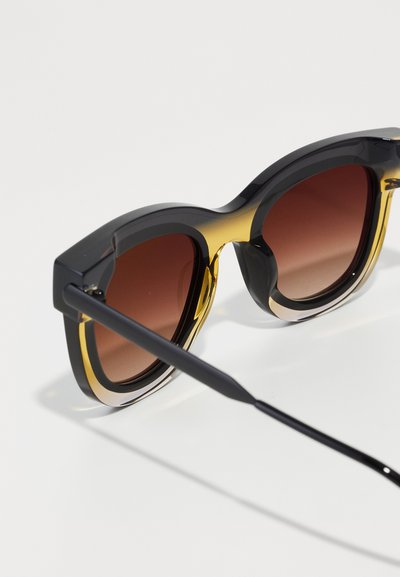 Melna un dzeltena acetāta saulesbrilles ar apaļām kārbām, pakāpeniski brūnām lēcām un elegantām melnām koriģējošām kājām. Malu apdare uzlabo dizainu.