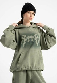 Młoda osoba w oliwkowym, oversized hoodie i dopasowanych spodniach, dostosowująca kołnierz, nosząca czarną wełnianą czapkę z metalowymi pierścieniami.