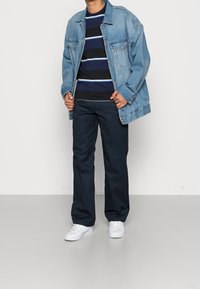 Jeansjacka över en marinblå och blå randig långärmad skjorta, kombinerad med marinblå byxor och vita sneakers. Avslappnad, lös passform.