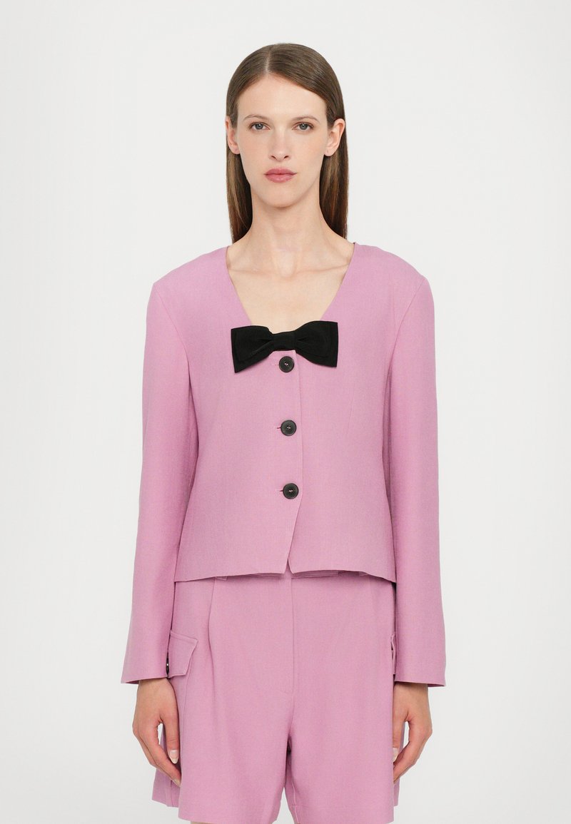 Light pink cropped jacket with a v-neck, black bow detail at the neckline, and front buttons. Soft fabric, long sleeves, and tailored fit.