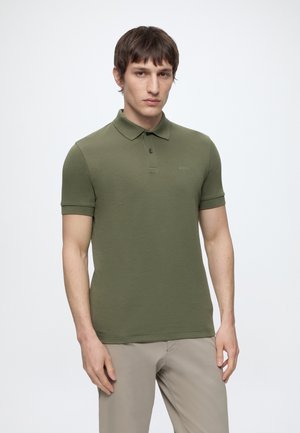 Jeune homme portant un polo vert olive à manches courtes et un pantalon beige, debout devant un fond clair uni, face à l'avant.
