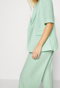 Blazer vert menthe à manches courtes et à revers cranté, avec une fermeture à un bouton et des poches latérales, associé à un pantalon assorti.