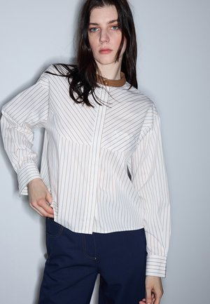 Frau mit langen dunklen Haaren, die eine weiße Bluse mit dünnen braunen Streifen und dunkelblaue Hosen trägt, steht vor einem schlichten hellen Hintergrund.