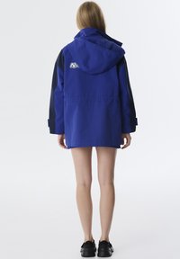 Blaue Kapuzenjacke mit schwarzen Ärmeln, ausgestattet mit einem Logo auf dem Rücken, sichtbaren Nähten und einem lässigen Schnitt. Strapazierfähiger Stoff mit glatter Textur.