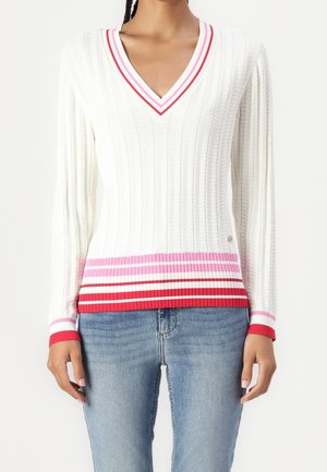 Vrouw draagt een witte kabelgebreide V-hals trui met rood, roze en wit gestreepte boord, gecombineerd met een blauwe spijkerbroek.
