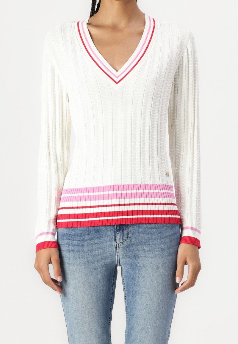 Femme portant un pull blanc torsadé à col en V avec une bordure rayée rouge, rose et blanche, associé à un jean bleu.