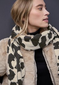 Écharpe beige et verte foncée à motifs en matériau doux, enroulée autour du cou, associée à une veste en fourrure beige et un haut noir.