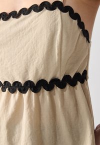 Beige kjole med strukturert stoff og svart scalloped kant på halslinjen og midjen, som skaper et kontrasterende designelement.