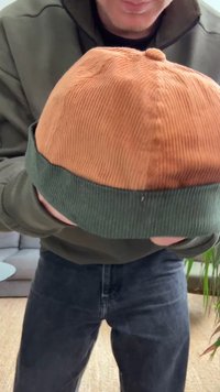 Kordurový klobúk typu bucket so striebristo-oranžovým vrchom a tmavozeleným rebrovaným okrajom. Textúra je mäkká s výraznými vertikálnymi ryhami.