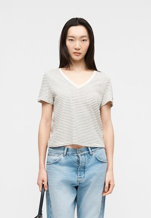 Junge Frau, die ein weißes, gestreiftes T-Shirt mit V-Ausschnitt und hellblaue Jeans trägt und eine dunkle Handtasche in der linken Hand hält.