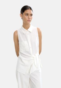 Biała bezrękawnik wykonana z lekkiego materiału, z koronkową kieszenią, kołnierzykiem i wiązaniem w talii.