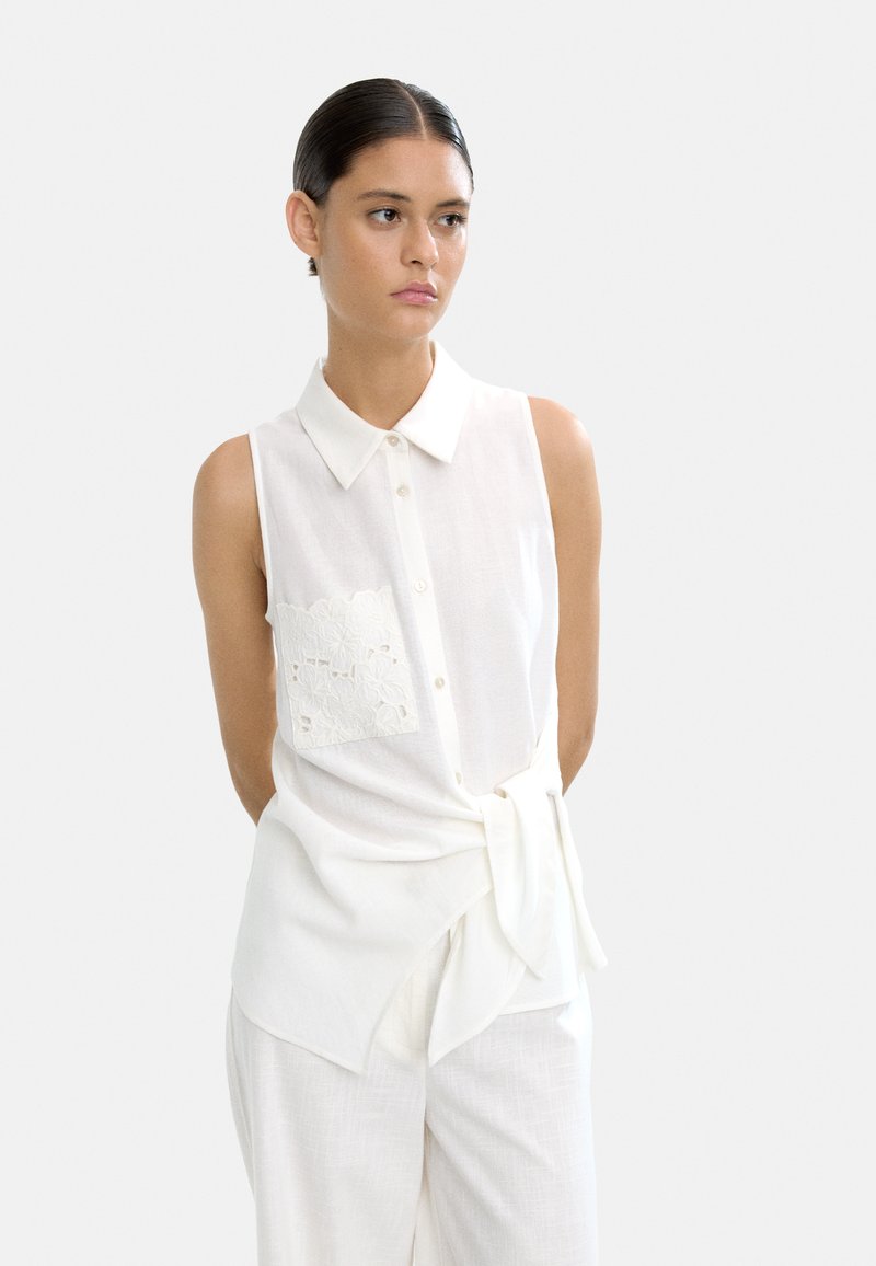 Biała bezrękawnik wykonana z lekkiego materiału, z koronkową kieszenią, kołnierzykiem i wiązaniem w talii.