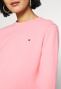 Frau mit kurzem schwarzen Haar, die goldene Kreolen trägt und ein pinkes Sweatshirt mit einem kleinen rot-weißen und schwarzen Logo auf der Brust anhat.