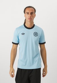 Lyseblå atletisk trøje med sort krave og manchetter. Har tekstureret stof og et sort cirkulært logo på brystet. Tre-stripes logo på skulderen.