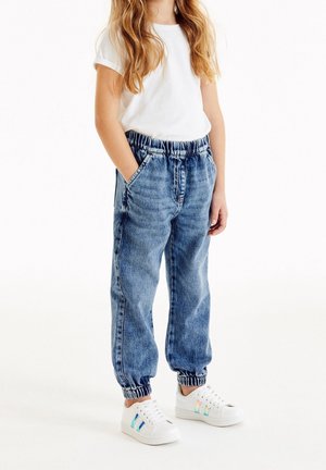 Kind draagt blauwe spijkerbroek met elastische tailleband, wit T-shirt met korte mouwen en witte sneakers met iriserende zijstrepen.