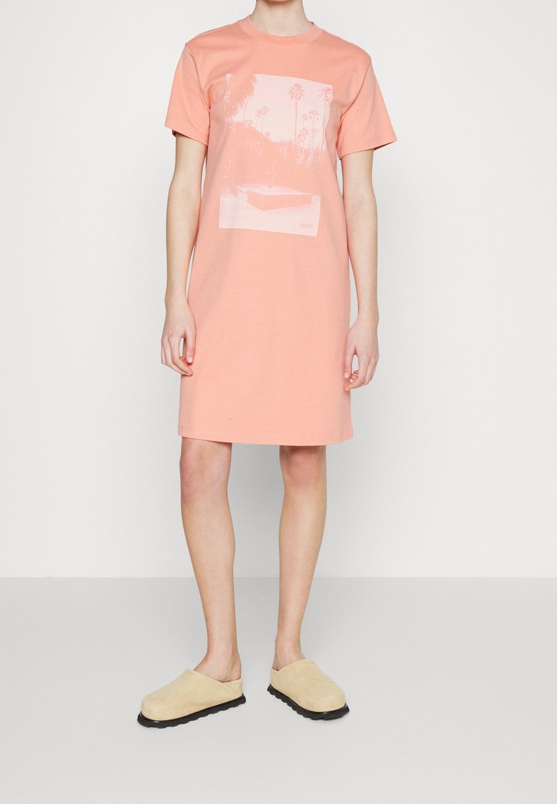 Robe t-shirt couleur pêche claire en coton, avec un motif graphique frontal de palmiers et une piscine, manches courtes et coupe droite.