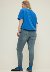 Camiseta de algodón azul con mangas cortas, combinada con jeans ajustados de un azul claro. Calzado negro tipo slip-on y calcetines azules visibles, fondo neutro.