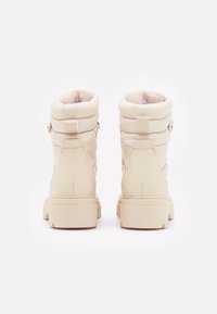 Bottines rembourrées beiges avec une finition en cuir texturé, bout arrondi et semelle en caoutchouc plate. Dotées d'une doublure en fausse fourrure et d'accents de boucle sur le côté.
