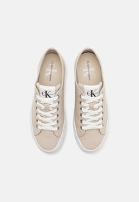 Beige Leinenschuhe mit weißen Gummisohlen, ausgestattet mit weißen Schnürsenkeln und einem dezenten Logo auf der Zunge. Flaches, rundes Design und glatte Textur.