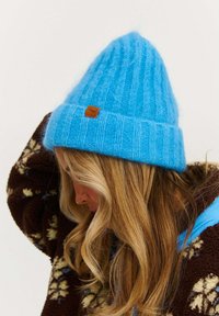 Modrá pletená čiapka so žebrovanou textúrou a malou hnedou koženou štítkou. Čiapka má špicatú korunku a široký preložený lem.