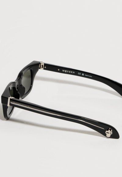Μαύρα γυαλιά ηλίου σχεδιαστή με χοντρές βραχίονες, ασημένια λεπτομέρεια κρανίου σε έναν βραχίονα και τη σήμανση "McQueen" εσωτερικά στο πλαίσιο.