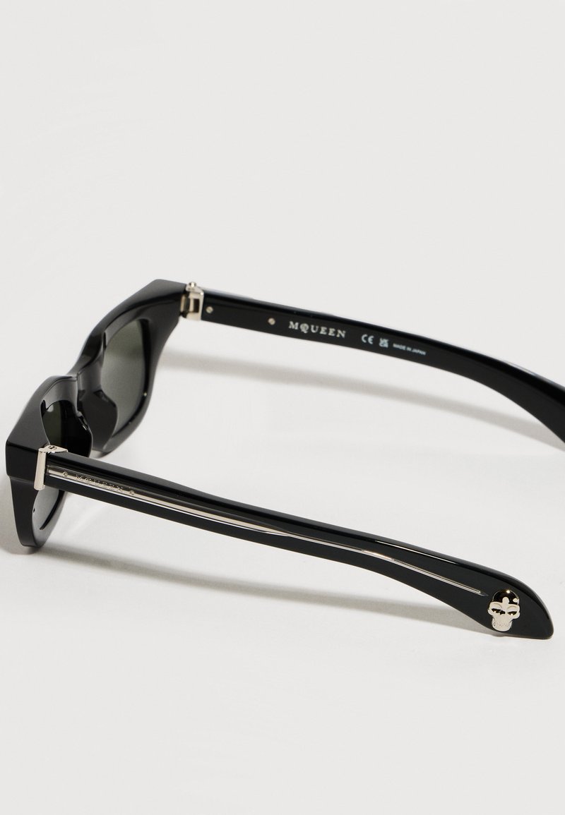 Occhiali da sole da designer neri con aste spesse, dettaglio teschio argentato su un'asta e marchio "McQueen" all'interno della montatura.