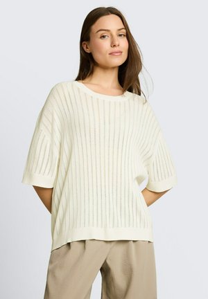 Kobieta z długimi brązowymi włosami, ubrana w luźny biały dzianinowy sweter i beżowe spodnie, stojąca na jednolitym jasnym tle.