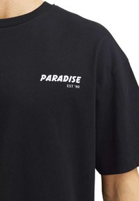 T-shirt nera in cotone con maniche corte e colletto tondo. Presenta sulla parte frontale, a sinistra, una grafica bianca con il testo "PARADISE EST '90".