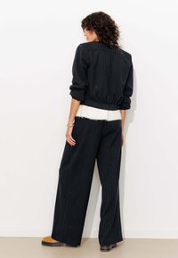 Veste oversized noire à rayures fines avec des poignets côtelés, associée à un pantalon à jambes larges et une ceinture douce crème. Texture décontractée et coupe ample.