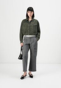 En kvinne med rett, mørkt hår har på seg en kort grønn jakke, grå cargo-bukser, svarte slingback-sandaler, og holder en liten svart håndveske.