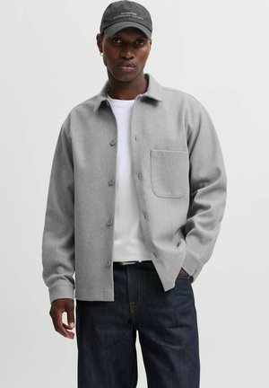 Homme portant une veste grise boutonnée sur une chemise blanche, un jean foncé et une casquette noire avec du texte, debout devant un fond clair uni.