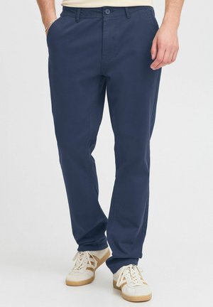Mann trägt marineblaue Freizeithose und weiße Sneakers vor schlichtem hellem Hintergrund, eine Hand in der Tasche, die andere entspannt.