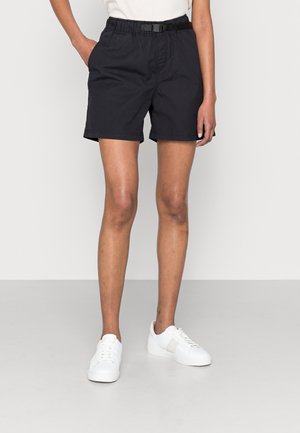 Personne portant un short noir décontracté avec une taille élastique et une boucle noire, associée à des baskets blanches, debout devant un fond uni.