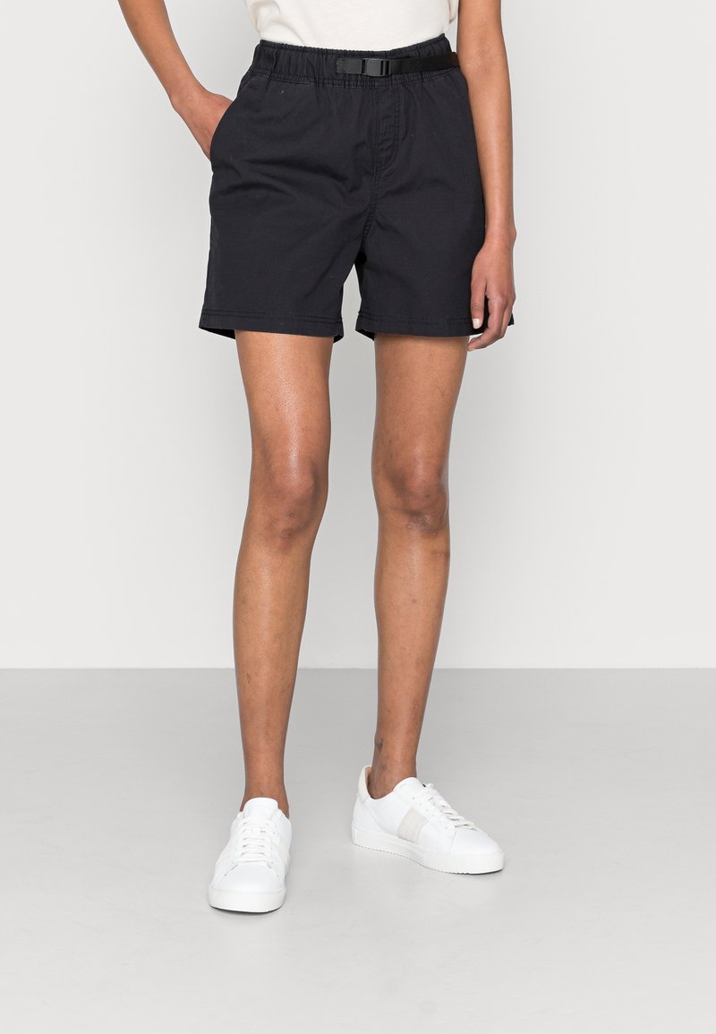 Personne portant un short noir décontracté avec une taille élastique et une boucle noire, associée à des baskets blanches, debout devant un fond uni.