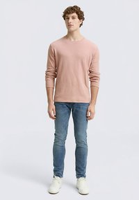 Chemise à manches longues rose clair en matériau doux, associée à un jean bleu et des baskets blanches. Design simple et ajusté avec un col rond.