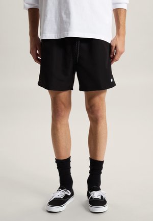 Uomo con pantaloncini neri, calzini neri, scarpe da ginnastica bianche e nere e maglia a maniche lunghe bianca, in piedi contro uno sfondo uniforme.