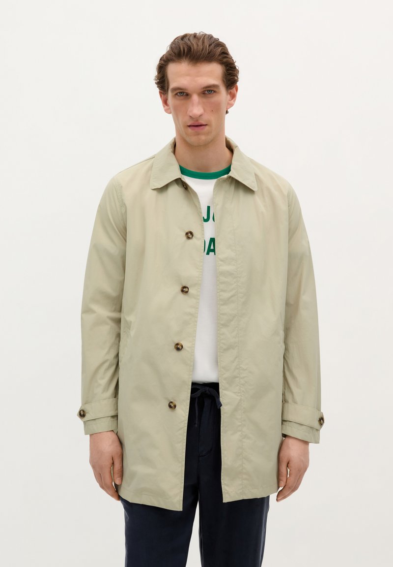 Khaki lehký trench coat s knoflíkovým zapínáním, špičatým límečkem a srolovanými rukávy, vrstvený přes bílou košili se zeleným lemováním.
