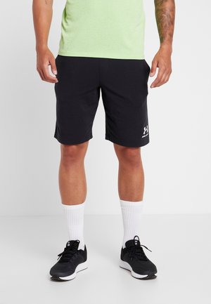 Zwarte sportshorts gemaakt van lichtgewicht stof, met een gladde textuur en een wit logo op de linkeronderkant. Gecombineerd met witte sokken en zwarte sneakers.