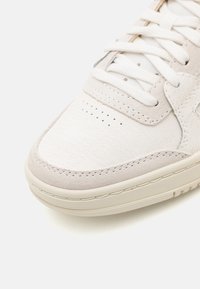 Beige sneakers med texturerat mockaskinn, vit snörning och perforerat tådetalj. Utrustad med en krämfärgad gummisula för stabilitet.