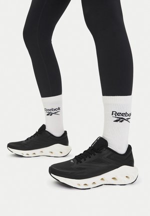 Czarne sneakersy Reebok z teksturowaną cholewką, białą podeszwą EVA i czarnymi sznurowadłami. W połączeniu z białymi skarpetkami z czarnym logo.