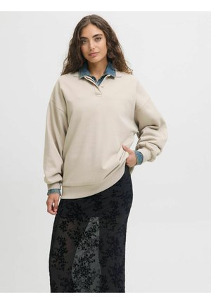 Pull beige avec un col en denim, doté d'une patte de boutonnage et de manches longues, associé à une jupe noire transparente ornée de motifs floraux.