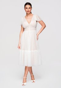 Femme portant une robe midi blanche transparente à pois avec des manches volantées et un col en V, associée à des escarpins blancs à bride cheville.