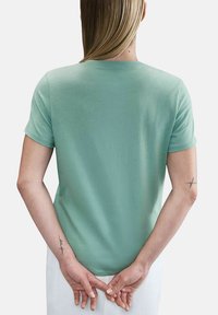 Personne avec des cheveux blonds portant un t-shirt à manches courtes vert clair et un pantalon blanc, les mains croisées derrière le dos, des tatouages visibles sur les avant-bras.