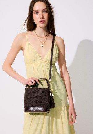 Jeune femme en robe jaune clair sans manches tenant un sac à main tressé marron foncé avec des détails en cuir et un pompon.